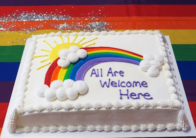 White cake with rainbow, sun, clouds, and All Are Welcome Here message.