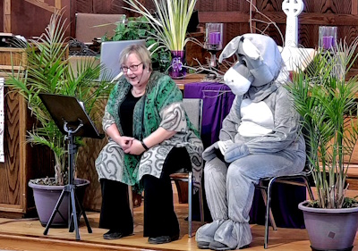 Rev. Cathy seated with person dressed as a donkey on the chancel talking to people out of picture during Sunday worship.