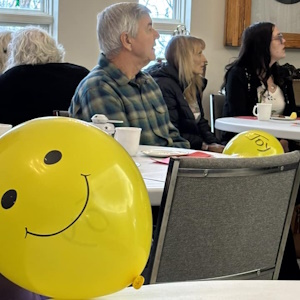 Tables with people seated in the background and a yellow balloon with a big smiley face in the foreground.
