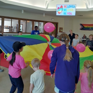 Children holding a colourful nylon fabric around the edge in mid-air with balloons above in the BUC sanctuary